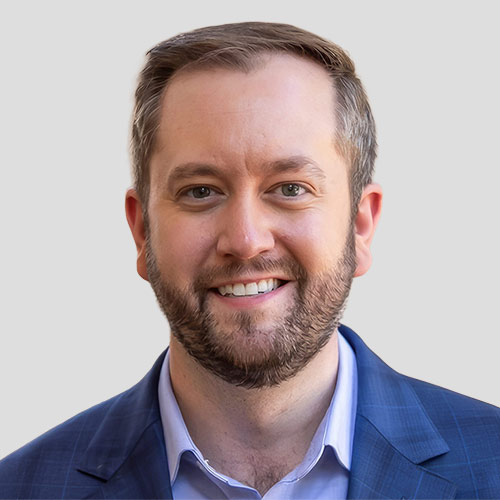 Professional headshot: smiling man in a blue suit jacket and light shirt against a neutral background.