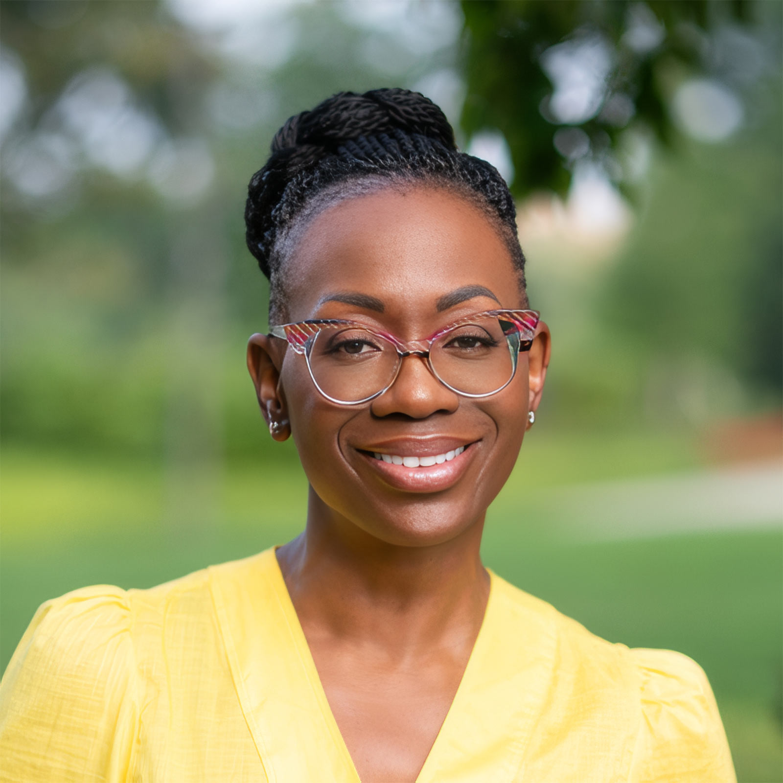 Close-up portrait of a smiling woman with braided bun, colorful glasses, and a yellow top outdoors.