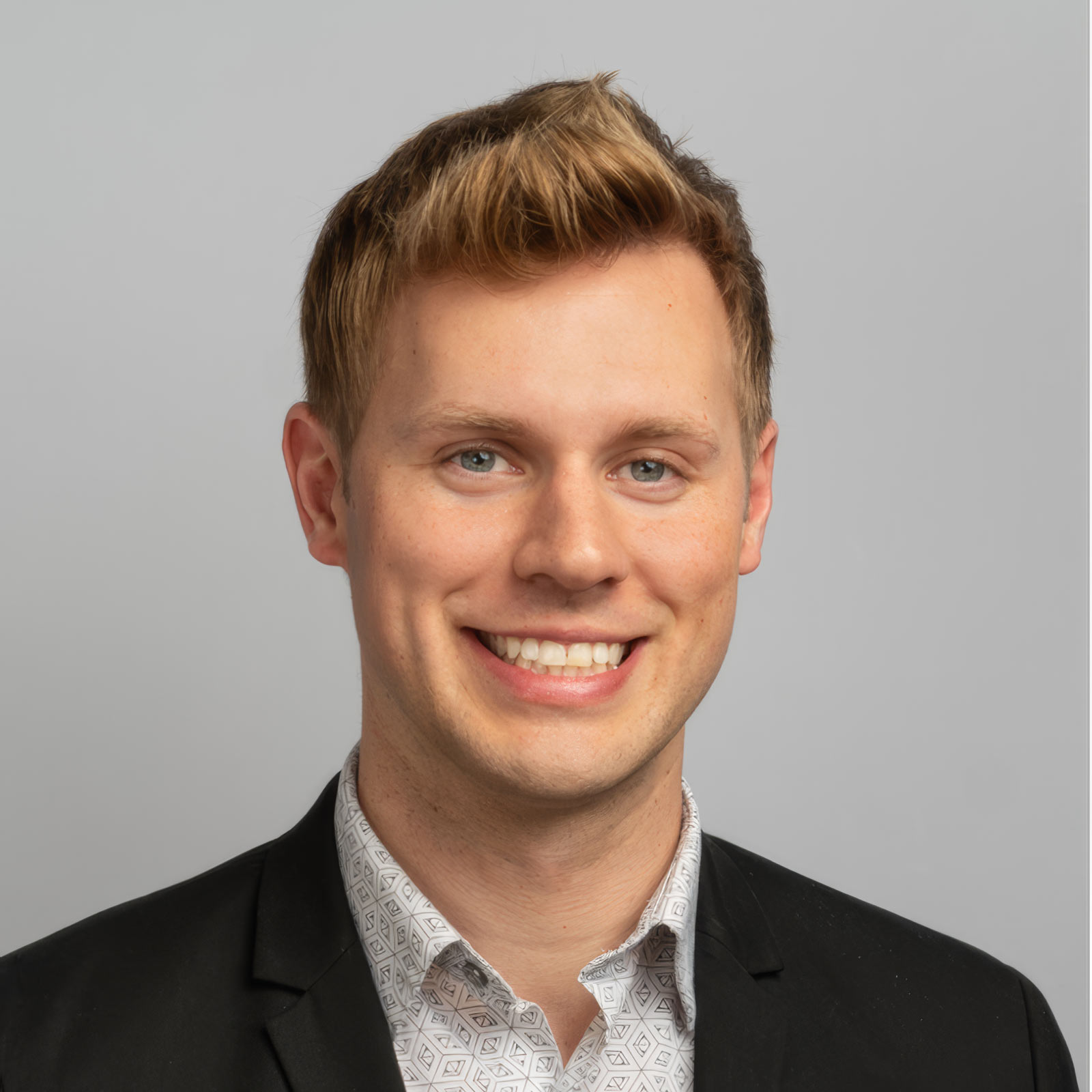 Smiling young man in a black blazer and patterned shirt, posing for a head-and-shoulders portrait against a gray backdrop.